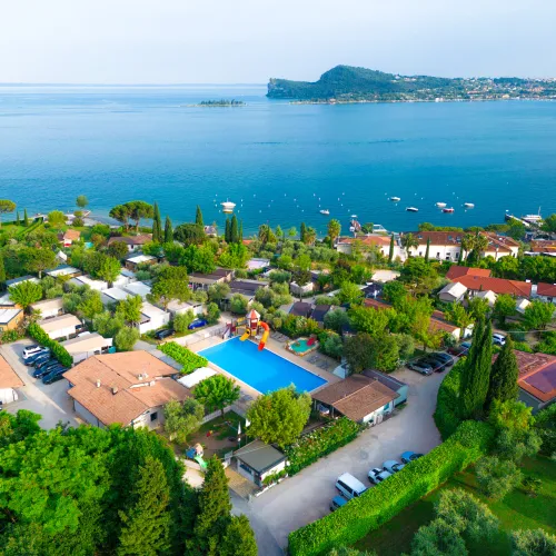 Aerial view of a lakeside resort at lake garda with pool, trees, and distant island.
