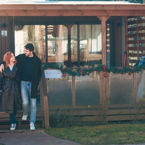 Lago Idro Winter Glamping accommodations.jpg Couple smiling in front of a wooden cabin decorated with garlands.