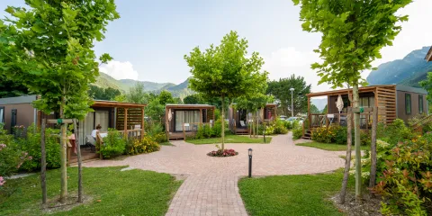 Pathway leading through a green park with cabins and trees against a mountain backdrop.