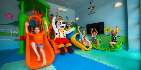 Children playing on colorful slides with a mascot in a playful, indoor setting.
