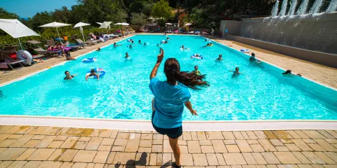 Person jumping into a crowded swimming pool on a sunny day, surrounded by trees.