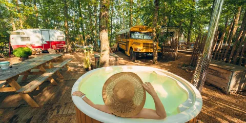 Person in a hot tub, wearing a straw hat, in a forest campsite with a bus and caravan.
