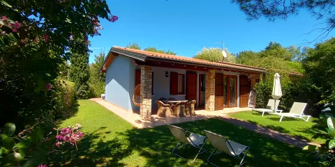Small house with terrace in a sunny garden, surrounded by trees and flowers.