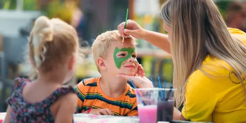 Face painting at a table, child with green mask, colorful cups.