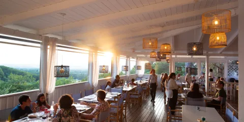 Sunlit restaurant with people dining, wooden interior, scenic view outside.