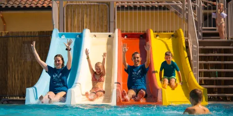 Four people joyfully sliding into a pool from colorful slides under a sunny sky.