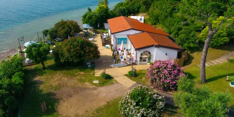 Coastal house with red roof, surrounded by trees and flowers, near blue water.