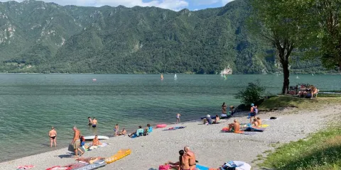 Beachgoers on a pebbly shore with green mountains in the background.
