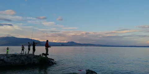 People fishing on a rock by a calm lake at sunset.