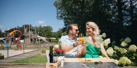 Couple toasting drinks at a park picnic table, sunny day.