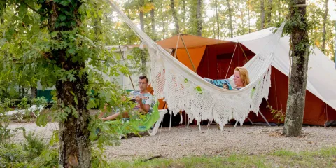 Two people relaxing in a hammock near a tent in a forest campsite.