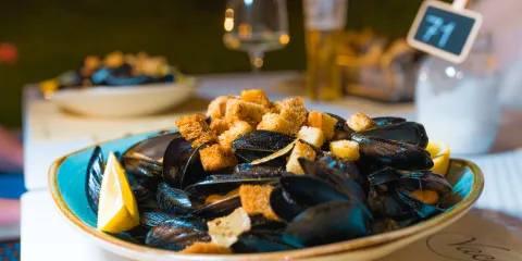 Bowl of mussels with croutons and lemon slices on a table, glassware in the background.