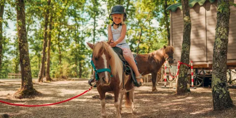 Child riding a pony in a forested area, wearing a helmet.