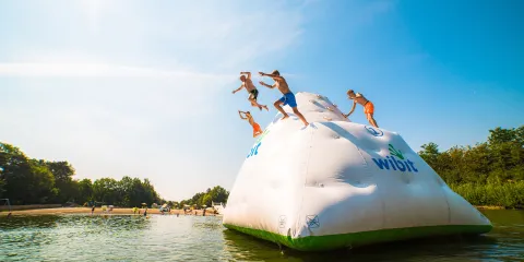 Kids jumping off large inflatable iceberg into water, sunny day.