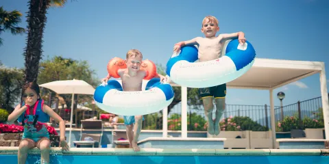 Children jumping into a pool with floatation rings, under a clear blue sky.