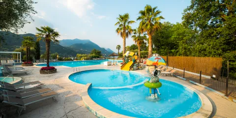 Sunny day at a swimming pool with palm trees, mountains, and a waterslide.