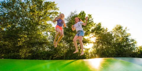 Kids jumping on a green inflatable, trees behind, sunlight peeking through.