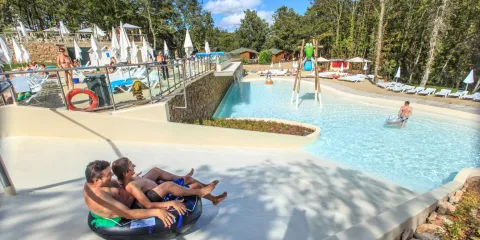 Two people on a waterslide in a sunny outdoor pool area with trees.