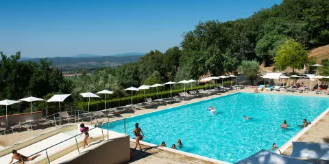Outdoor pool with people swimming, surrounded by trees and distant hills under a clear blue sky.