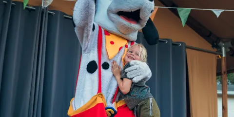 A child hugs a large dog mascot on stage under colorful bunting.