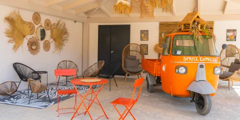 Orange vintage truck in a cozy patio with wicker decor and red metal chairs.