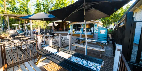 Outdoor cafe with umbrellas, wooden tables, and trees in the background.