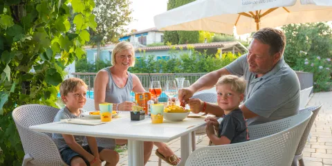 Family dining outdoors under a large umbrella on a sunny day.