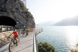Cyclists ride on a lakefront path near a mountain tunnel at sunset.