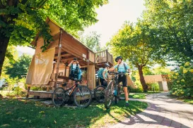Cyclists resting by a wooden cabin, surrounded by trees on a sunny day.