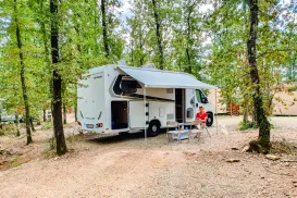 Camper van parked in a wooded campsite with chairs and table outside.