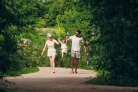 Family walking on a path in a lush, green park, lifting a child between them.