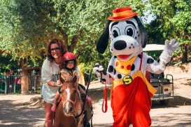 Child rides a pony with a woman; Spotty waves nearby.