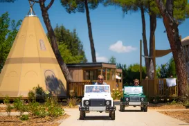 Two people driving mini cars near a large teepee-style tent.