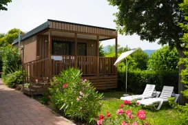 Cozy wooden cabin with a porch, surrounded by greenery and flowers; two lounge chairs nearby.
