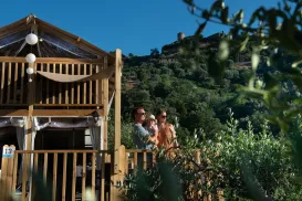 Wooden house with people on balcony, lush greenery, and hill in background under blue sky.