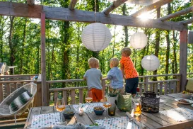 Children on a sunlit deck, surrounded by trees and lanterns, enjoying a late afternoon.
