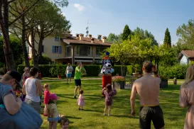 Adults and children gather on a grassy lawn, with trees and a house in the background.