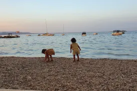 Two children play on a pebbled beach at sunset, boats in the background.