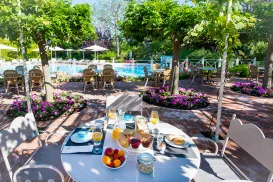 Breakfast table by a pool with trees and flowers nearby.