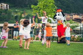 Children dancing with a cartoon dog mascot on a grassy lawn.