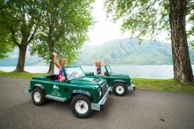 Two kids in toy cars waving by a lake, with mountains in the background.