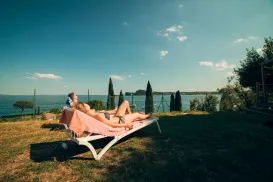 Two people relax on a lounger by a lake under a sunny blue sky.
