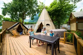 Family on a wooden deck by tents, surrounded by trees and summer greenery.