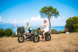 Two people riding bikes on a grassy hillside by a lake under a clear blue sky.