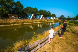 Father and child fishing by a pond, cabins in the background, sunny day.