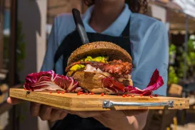 Waitress holding a gourmet burger with colorful garnishes on a wooden board.