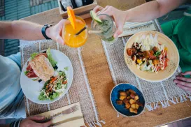 People toasting drinks over a table with sandwiches and salad.