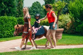 Child riding a small horse with two adults walking beside, in a garden setting.