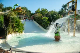 Water slide with animal statues and people splashing, surrounded by greenery.