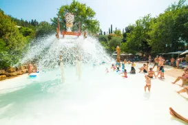 Children and adults play in a sunlit water park with falling water from a bucket.